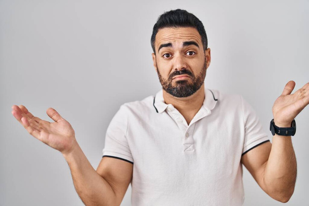 Young hispanic man with beard wearing casual clothes over white background clueless and confused expression with arms and hands raised. doubt concept.  - Foto, immagini