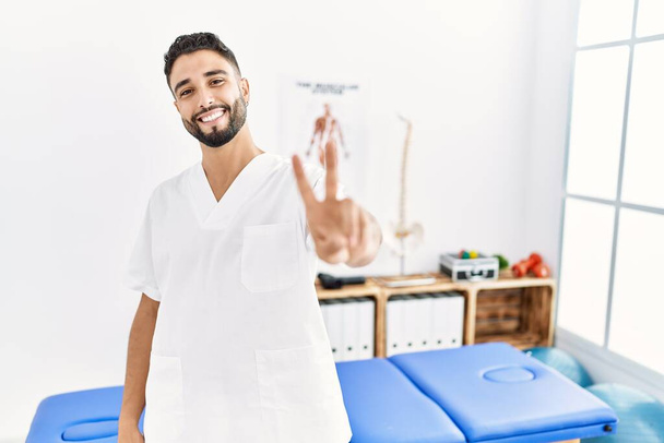 Ein junger gutaussehender Mann mit Bart, der in einer Schmerzklinik arbeitet, lächelt in die Kamera und zeigt Finger, die ein Siegeszeichen machen. Nummer zwei.  - Foto, Bild