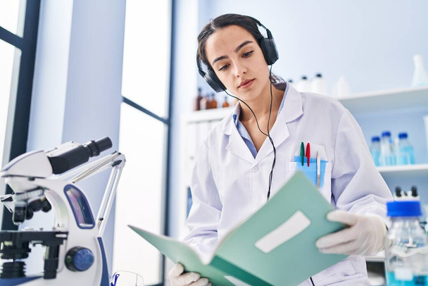 Young hispanic woman wearing scientist uniform listening to music reading book at laboratory - Fotoğraf, Görsel