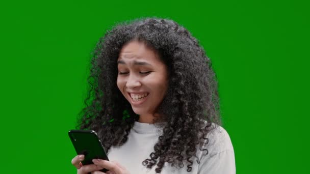 zadowolona kobieta z kręconymi długimi włosami ubrana w białą koszulę trzymając smartfona wpisując SMS z przyjacielem śmiejąc się Izolowane na zielonym ekranie - Materiał filmowy, wideo