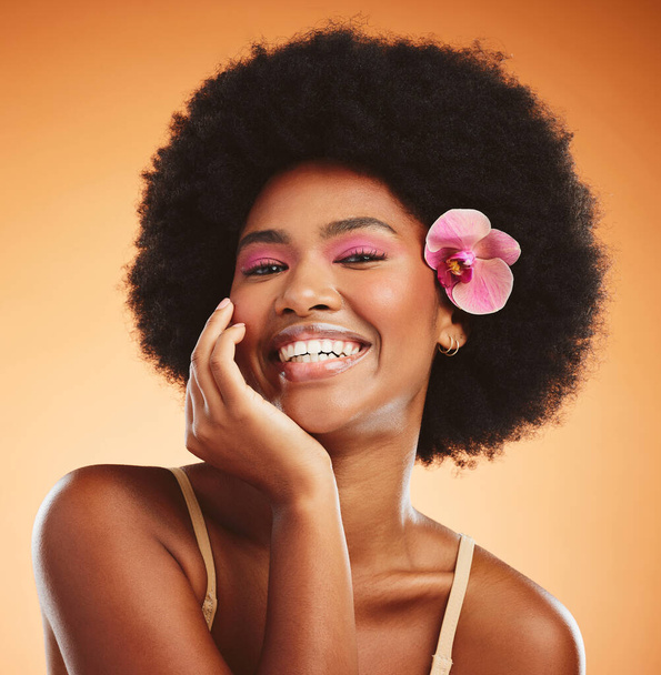 Face, beauty and black woman with flower in afro hair on orange studio background. Portrait, skincare and happy woman or female model from Nigeria in pink makeup, cosmetics and orchid in healthy hair. - Zdjęcie, obraz
