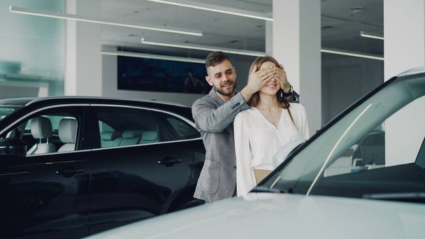 Cheerful bearded guy is making surprise for his wife closing her eyes with his hands and leading her to beautiful new car is motor showroom, happy woman is hugging and kissing him. - Fotografie, Obrázek