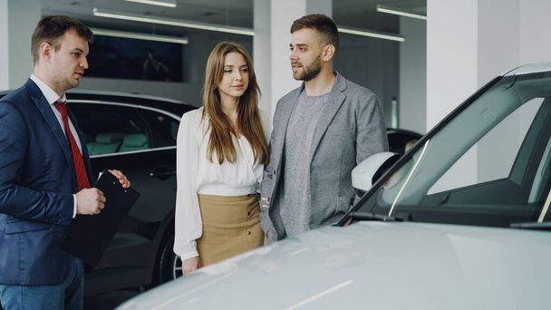 Friendly car salesman in suit is talking to confident young man telling him about new vehicle model while pretty woman is standing near her husband holding his arm and smiling. - Фото, зображення
