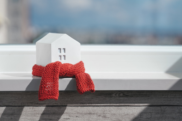 Miniatuur huis in een rode sjaal op de vensterbank. Het concept van passieve huisverwarming. Thermische isolatie van een gebouw of woning. Energiecrisis. - Foto, afbeelding
