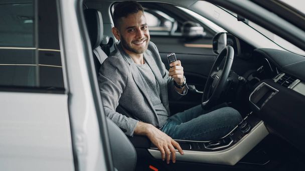 Close-up portrait of handsome bearded guy new car owner sitting inside beautiful automobile holding key fob and smiling looking at camera. Transportation and people concept. - Fotó, kép