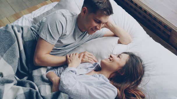 Cheerful caucasian man is lying in bed with his asian wife holding hands and talking laughing. relationship and love concept. - Fotografie, Obrázek