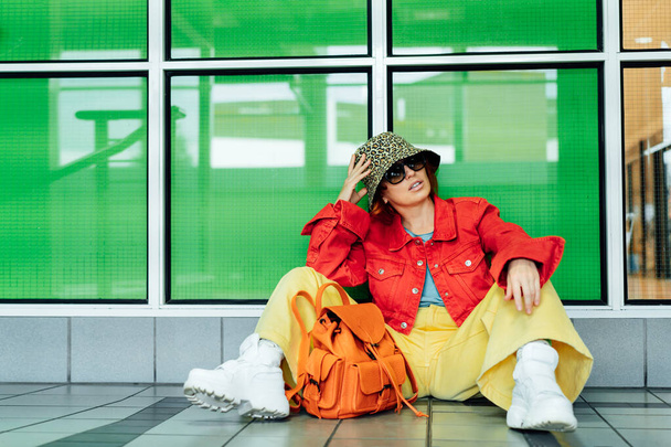 Hipster young woman in bright clothes, sun glasses, backpack bag and bucket hat posing while sitting near green glass wall background. Urban city street fashion. Fashion blogger. Selective focus - Фото, зображення