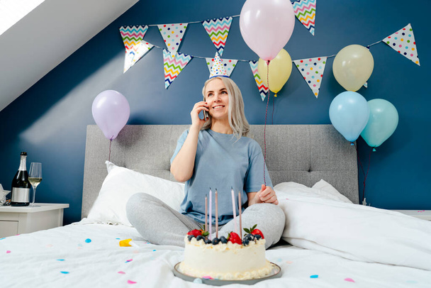 Happy emotional woman in pajama and party cap talking on phone at birthday morning while sitting on the bed with birthday cake and balloons in bedroom. Geting congratulations. Happy birthday concept. - Foto, afbeelding