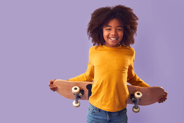 Studio Portrait Of Young Boy Holding Skateboard Behind Back Against Purple Background - 写真・画像