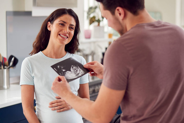 Excited Pregnant Transgender Couple At Home In Kitchen Looking At Ultrasound Scan Of Baby - Фото, изображение