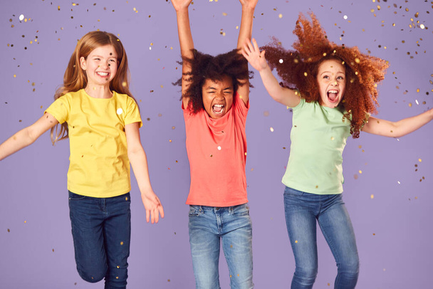 Studio Shot Of Children With Glitter Jumping In The Air With Outstretched Arms On Purple Background - Foto, Imagen