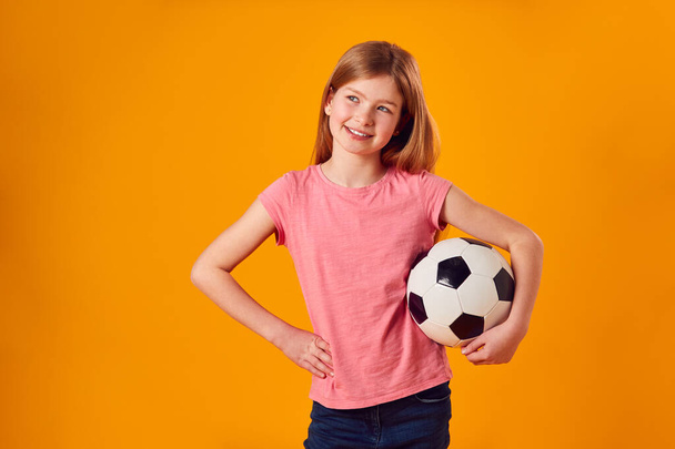 黄色の背景に腕の下でサッカーボールを保持している若い女の子のスタジオショット - 写真・画像