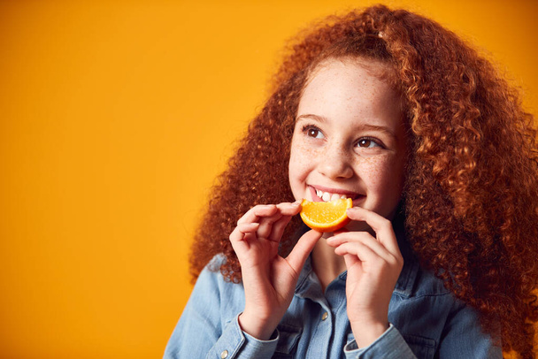 Studio Portrait Of Girl Holding Orange For Smiling Mouth Against Yellow Background - Valokuva, kuva
