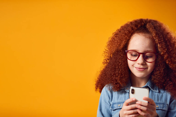 Stúdió portré fiatal lány viselő szemüveg segítségével mobiltelefon ellen sárga háttér - Fotó, kép