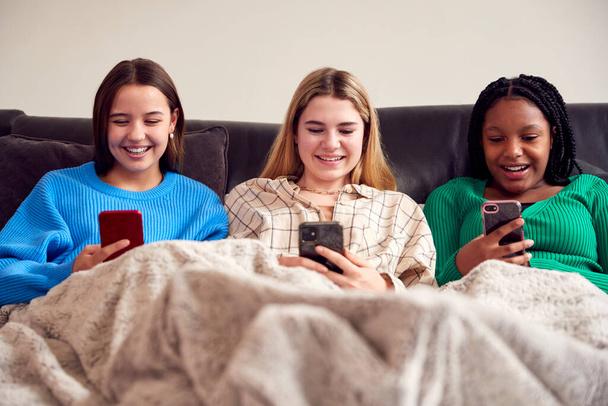 Group Of Multi-Cultural Teenage Girl Friends Snuggled Under Blanket Looking At Mobile Phones At Home - Foto, afbeelding