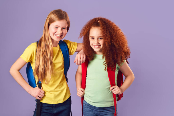 Studio Shot Of Two Girls With Backpacks Going To School On Purple Background - Фото, изображение
