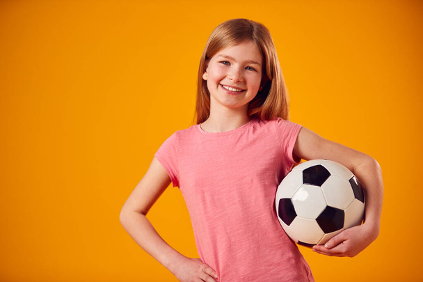 Studio Shot Of Young Girl Holding Soccer Ball Under Arm Against Yellow Background - Valokuva, kuva