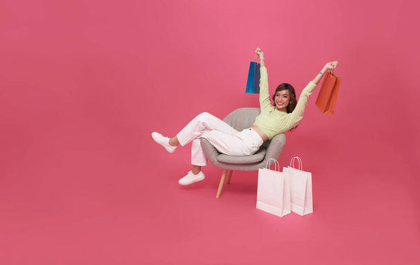 πορτρέτο του Happy Ασιάτισσα έφηβη γυναίκα κάθεται στον καναπέ με τσάντες ψώνια απομονωμένη σε ροζ φόντο, Shopper ή shopaholic έννοια. - Φωτογραφία, εικόνα