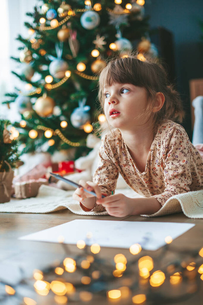 Dreaming cute little girl writing letter to Santa Claus or writing dreams of a gift with near Christmas tree. Merry Christmas and Happy New Year! - Foto, afbeelding