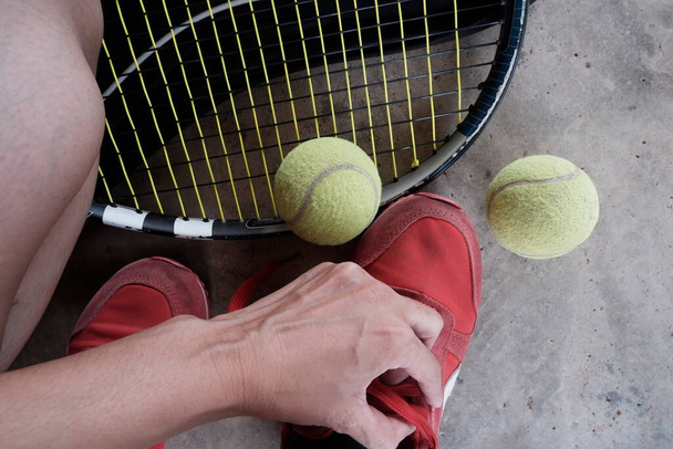 Μια παλιά μπάλα του τένις στο χέρι και μια ρακέτα του τένις σε μια τσάντα και παπούτσια στο πάτωμα. - Φωτογραφία, εικόνα