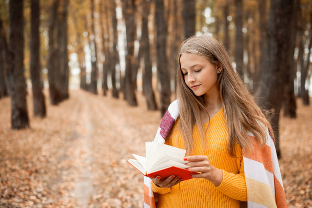 Красивая блондинка с книгой в руках в осеннем парке. Привлекательная девушка, читающая книгу в лесу во время прогулки - Фото, изображение