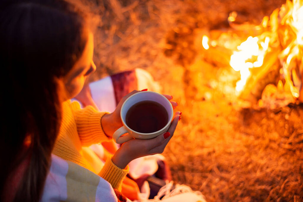 Beautiful girl drinking tea near the fire. Lonely girl with a cup of warm tea wrapped in warm plaid thinking about events, future, resting on nature - Photo, Image