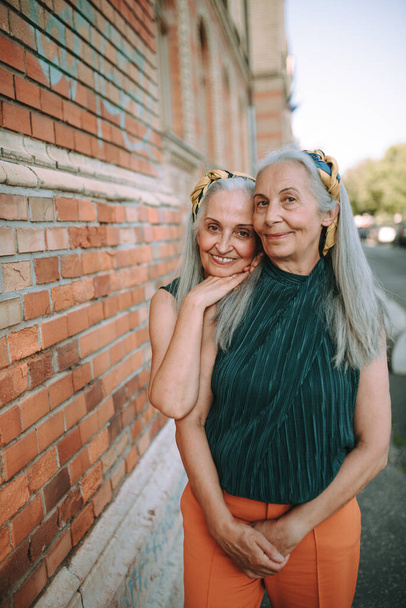 Donne anziane gemelle, con gli stessi vestiti in piedi e in posa davanti a un muro di mattoni, all'aperto in città. - Foto, immagini