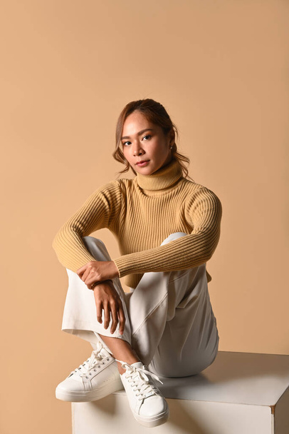 Elegant woman in stylish warm clothes sitting over beige background . Fashion studio photo, Autumn and Winter concept. - Fotó, kép