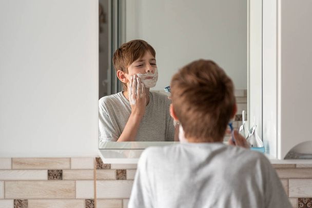 Little brunette boy applies shaving foam to face. Reflection in the mirror. Daily morning procedures. - Φωτογραφία, εικόνα
