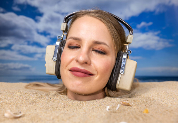 Una mujer bonita enterrada en su cabeza en una playa escuchando música con auriculares - Foto, imagen