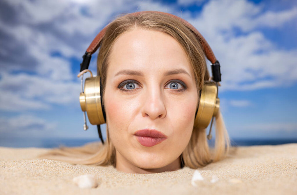 Μια όμορφη γυναίκα θάφτηκε μέχρι το κεφάλι σε μια παραλία ακούγοντας μουσική με ακουστικά. - Φωτογραφία, εικόνα