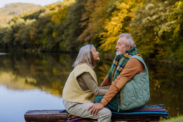 Senior couple in love sitting together on a bench near lake, during autumn day. - Foto, Imagem