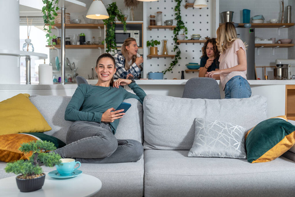 Young happy woman isolated herself sitting on sofa smiling while her friends standing in the background talking and gossiping their boyfriends. - Foto, Bild