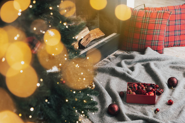 Caja de bolas de Navidad puesta en alfombra gris o cuadros listos para su uso para decorar y colgar en abeto verde de Navidad con luces, guirnalda en la sala de estar cerca de la chimenea con madera en el interior - Foto, imagen