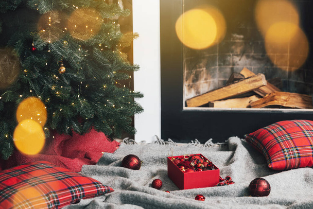 Doos met kerstballen liggend op grijs tapijt of plaid klaar voor gebruik voor decoratie en opknoping op groene kerstboom met verlichting, slinger in de woonkamer in de buurt van open haard met hout binnen - Foto, afbeelding
