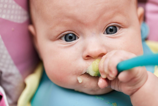 cute baby girl eating with spoon on baby chair in kitchen - Foto, Imagem