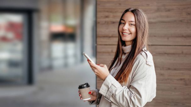 Nuori nainen käyttää matkapuhelinta hymyillen ja katsoen kameraan ja seisoen lähellä toimistorakennusta. Tyttö opiskelija seisoo lähellä yliopistoa. Internet-banneri. kopioi tila - Valokuva, kuva