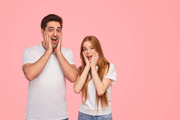 Astonished young boyfriend and girlfriend in white t shirts touching cheeks and looking at camera with opened mouth against pink background - Photo, image