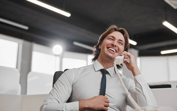 Low angle of cheerful man in white shirt adjusting tie and laughing while answering phone call from client during work in modern office - Foto, Bild