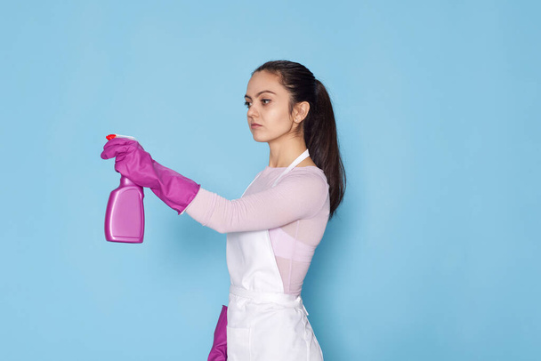 confident woman in purple rubber gloves and cleaner apron cleaning with detergent sprayer on blue background. side view - Foto, Imagem