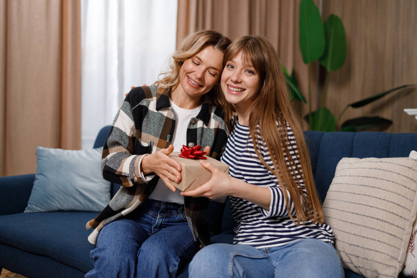 Dospělá žena dělá nečekané překvapení pro její matku středního věku, dává jí dárkovou krabici, zatímco sedí na pohovce v bytě - Fotografie, Obrázek