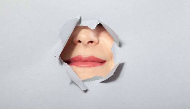 Surprised woman looking playfully in torn paper hole, has excited cheerful expression, looks through breakthrough of gray background. Wow, great news. Closeup - Foto, imagen