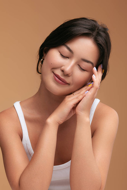 Glad young Asian female closing eyes and touching soft skin of face and hand during beauty routine against brown background - Φωτογραφία, εικόνα