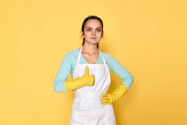 happy caucasian woman in rubber gloves and cleaner apron showing thumbs up gesture on yellow background. cleaning - Photo, Image