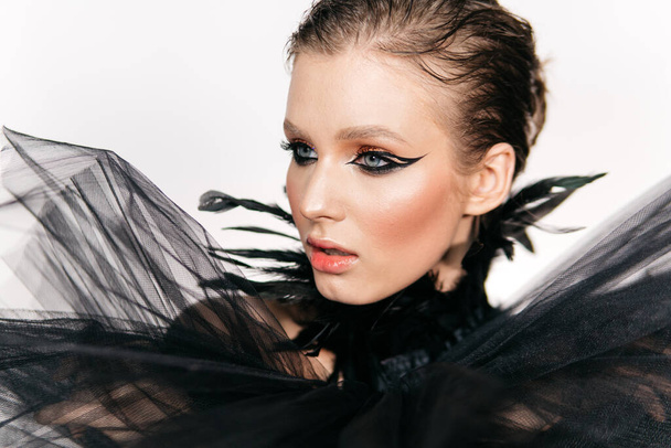 Hermosa chica con ojos ahumados maquillaje, pluma de plumas negras sobre un fondo blanco - Foto, Imagen