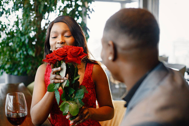 Een paar genieten van een dagje uit in het restaurant. Zwarte man schonk een boeket rode rozen aan een vrouw. Vrouw in rode elegante jurk en man blauw kostuum. - Foto, afbeelding