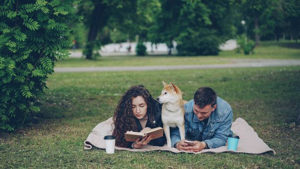 Young family spending weekend in the park, woman reading book, man using smartphone, cute dog resting between them. Guy is showing phone screen to his laughing wife. - Foto, Bild