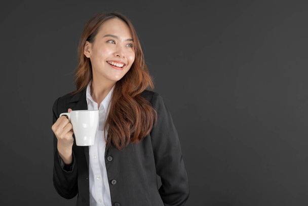 Mulher de negócios vestindo um terno preto de pé com uma xícara de café em um fundo preto. - Foto, Imagem
