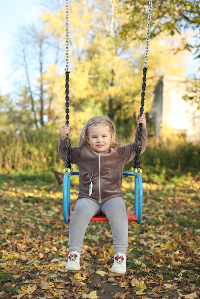 The child plays in the yard on the playground, rides on the swing, the girl is in a great mood, cheerful - Foto, Imagen