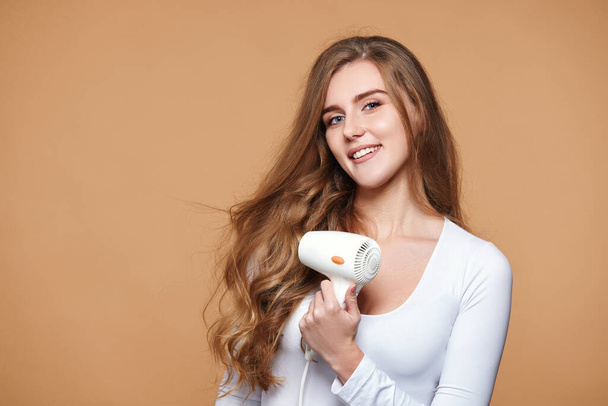 lovely woman with strong healthy curly hair using white hair dryer on beige background - Fotografie, Obrázek